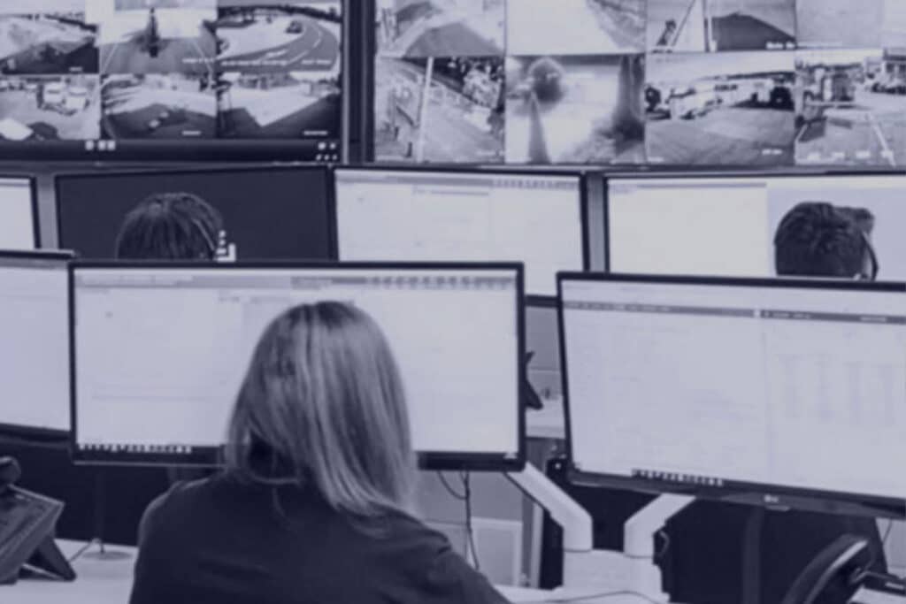 Security monitoring centre with operators at computer desks viewing multiple live CCTV feeds on large wall-mounted screens.